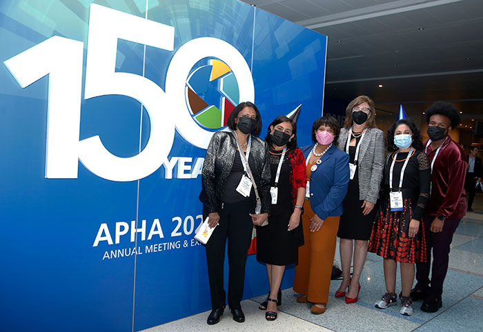 A group of six people wearing masks pose in front of an APHA 150th anniversary sign. 
