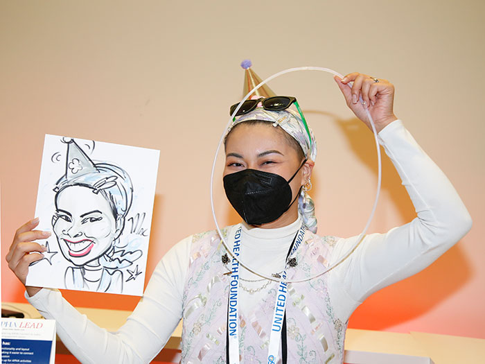 A young woman wearing a mask and a party hat holds up a caricature drawn of her in one hand and a small hoop in front of her face with the other. 