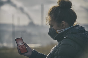 A woman check air pollution level on her cell phone, as smoke billow from factory in background