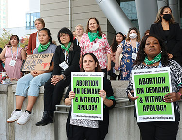 A group of women, looking serious, stand and sit on a wall at the reproductive rights rally. Some are holding signs that say “Abortion on demand and without apology” and “abortion is health care.” 