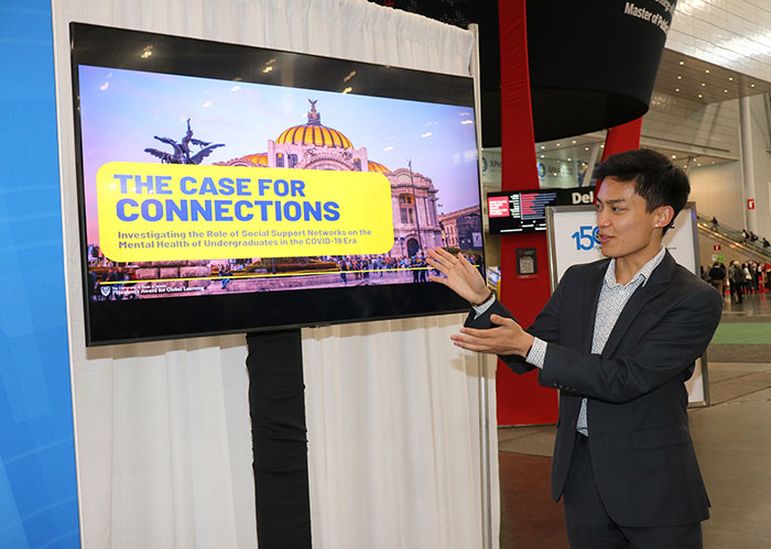 A young man in a black suit jacket motions to a digital screen next to him that says The Case for Connections. 