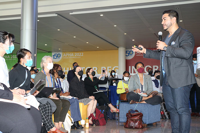 A man stands on the right in a sports jacket holding a microphone giving a presentation to a crowd sitting on benches, many with laptops or notebooks. 