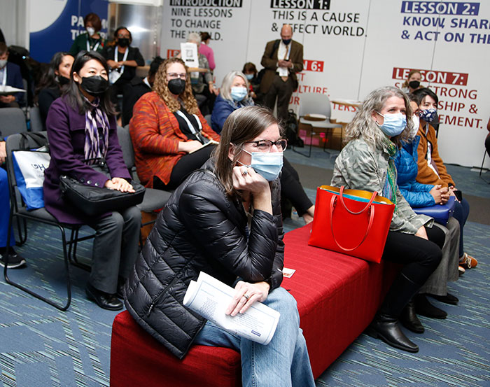 An attendee sits on a red bench in the expo hall theater lounge and leans forward intently with her head resting on her hand. 