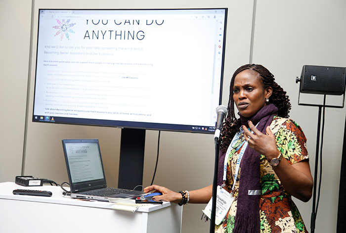 A woman stands in front of an audience with a digital screen behind her. 