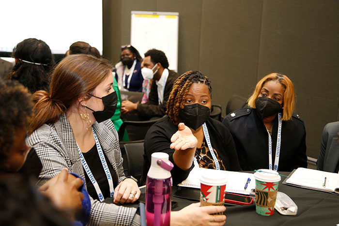 A woman in braids and a black mask emphasizes a point with a hand gesture while sitting at a table with other attendees. 