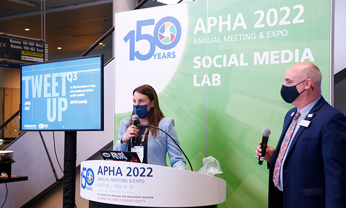 A woman stands in front of an APHA 2022 Social Media Lab sign with a microphone asking at the APHA 2022 Tweetup while a man stands off to the side with his own microphone. 