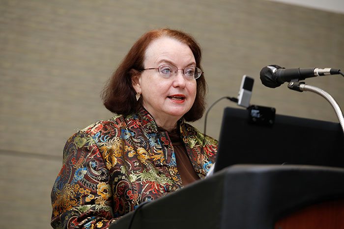 A person with red hair and a colorful jacket stands at the lectern to give a presentation. 