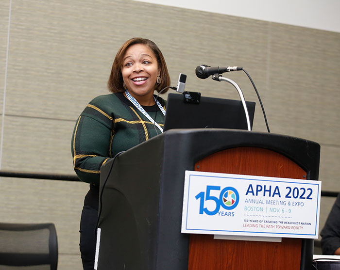 A person in a green sweater with gold stripes smiles at the audience as she stands at a lectern. 