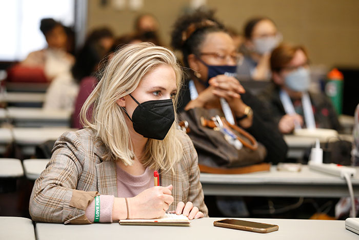 A blonde person wearing a black masks sits at a long table in a conference room and takes notes in a notebook. 