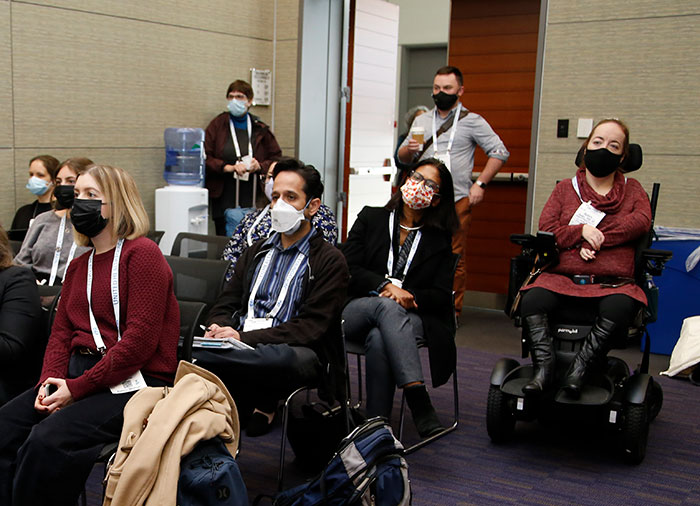 About seven people wearing masks sit in the back of a conference room listening to a speaker. 
