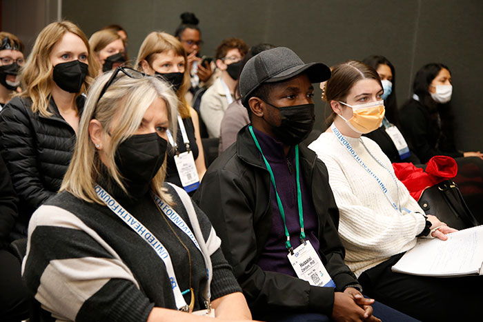 A group of people wearing masks sit in rows in the audience during a conference session. 