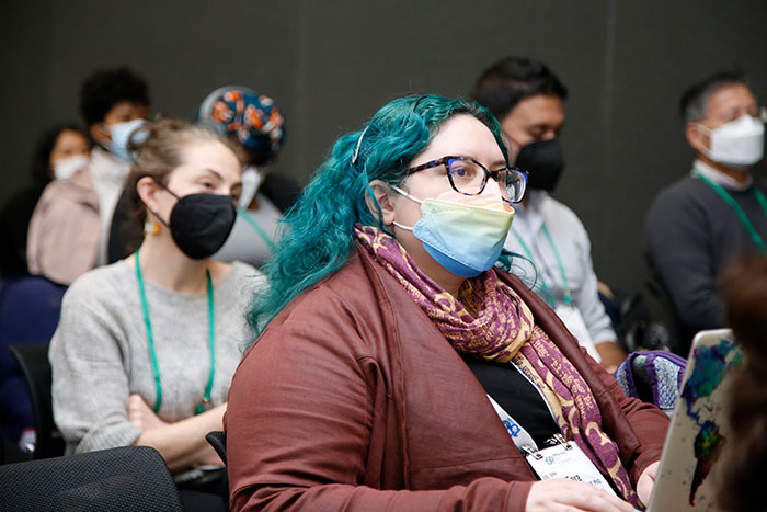 A person with dyed green hair and wearing a mask takes notes during a conference session. 