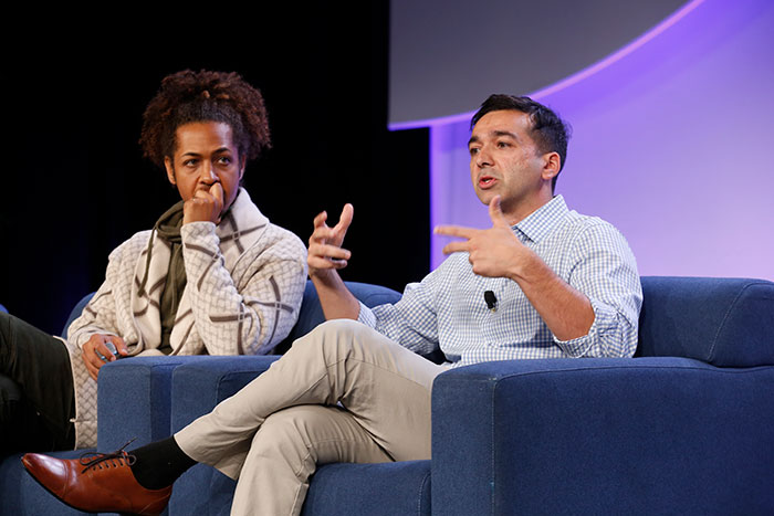 Two speakers sit in blue chairs on a large stage, as one talks during a presentation and other watches. 