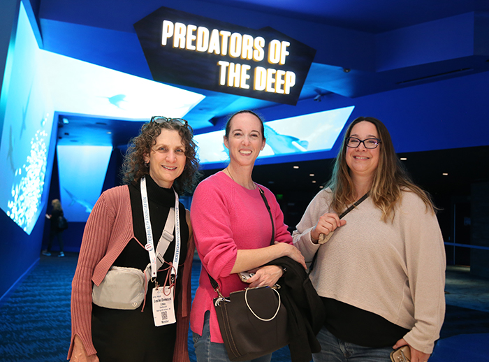 Three women stand together, smiling at the camera with an aquarium sign behind them and over their heads that reads Predators of the Deep.