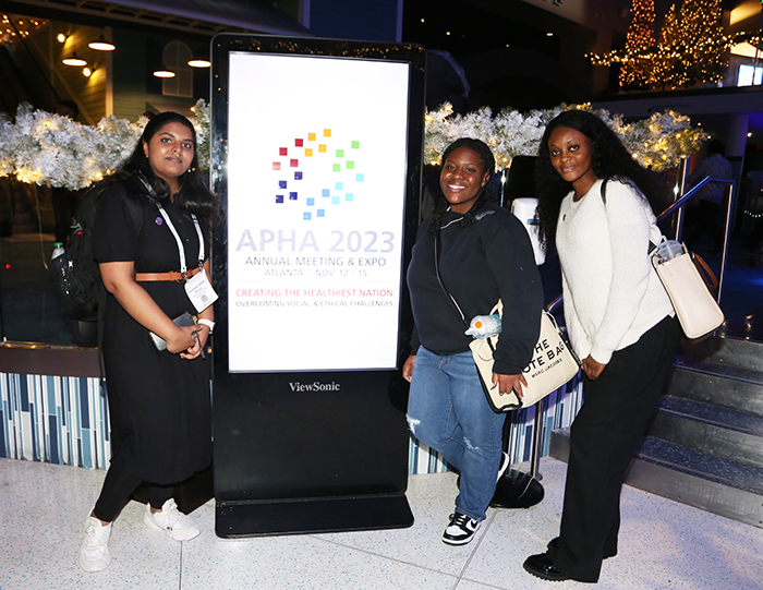 Three young adults pose for the camera as they stand on either side of a brightly lit APHA sign.