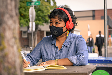 Close-up of a Filipinx woman with a filtering face mask, sitting at a table with notebook and pen. She has colorful flower earrings and headphones on while looking into the distance.