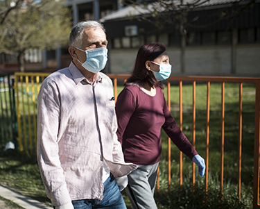 A man and woman, probably around age 65, wear casual clothes and surgical masks as they walk down a neighborhood street.