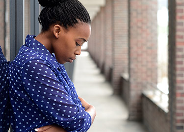 Woman stands outside leaning against wall with head down.