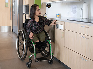 A South Asian person sits in her wheelchair and presses a button to lower the height-adjustable shelves in an accessible kitchen.