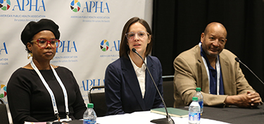 Three people on a panel sit at a table, while the person in the middle speaks into a microphone. 