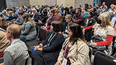 People sit next to each other in chairs in a crowded conference session room. 