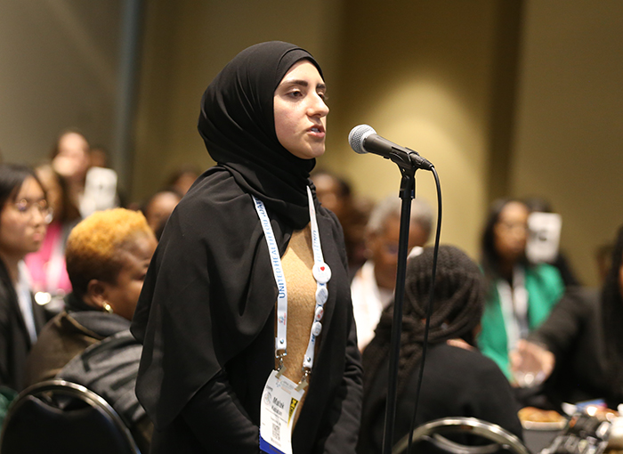 A young woman wearing a black hijab stands in front of a standing microphone and asks a question.