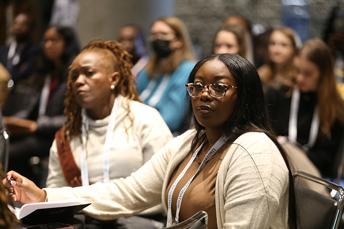 Two Black women sit in chairs in the audience listening to a presenter