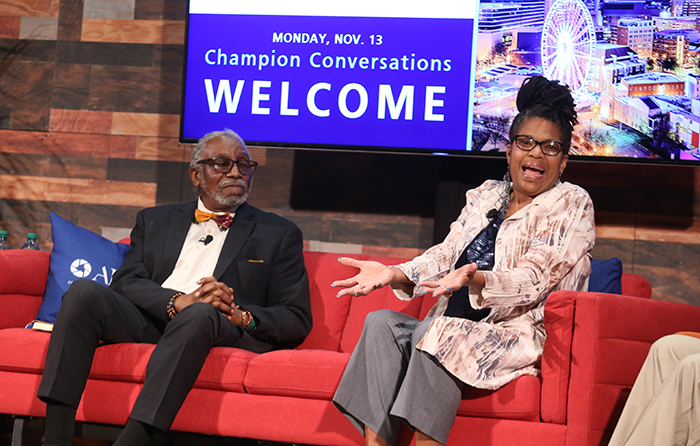 An older Black man in a black suit and red bow tie sits on a red couch next to a Black woman who is speaking to the crowd.
