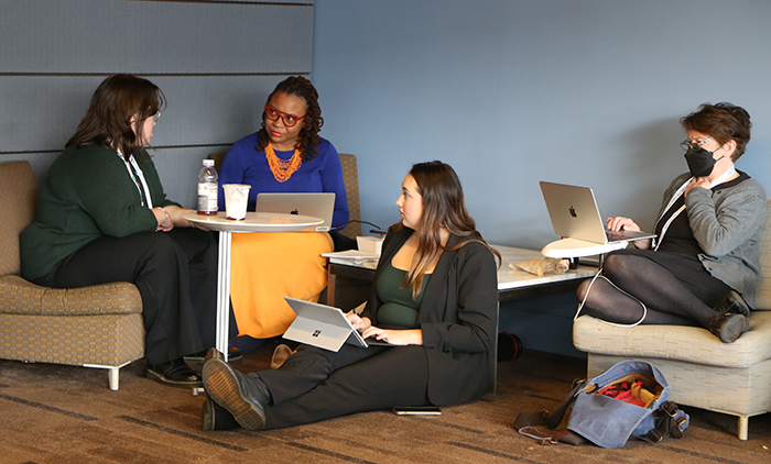 Four people gather together in chairs and on the floor talking and typing on laptops. 