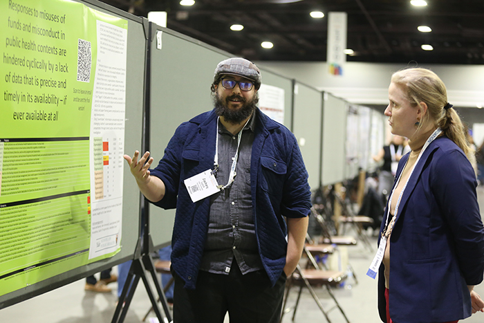 Man with beard and glasses and cap on his head talks about his poster to a woman.