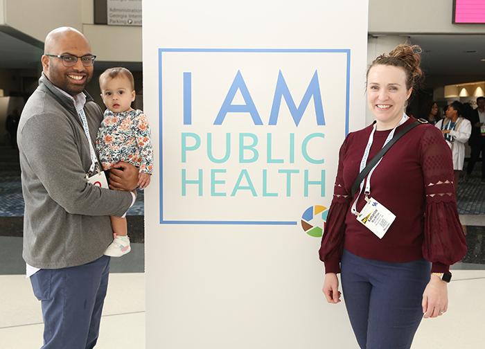 A Black man in a grey pullover and glasses and holding a baby poses on the left of an "I Am Public Health" sign, with a woman posing on the right of the sign. 