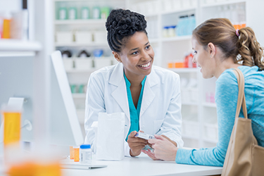 A Black woman pharmacist is leaning on a pharmacy counter and smiling at the white woman leaning on the counter next to her. 