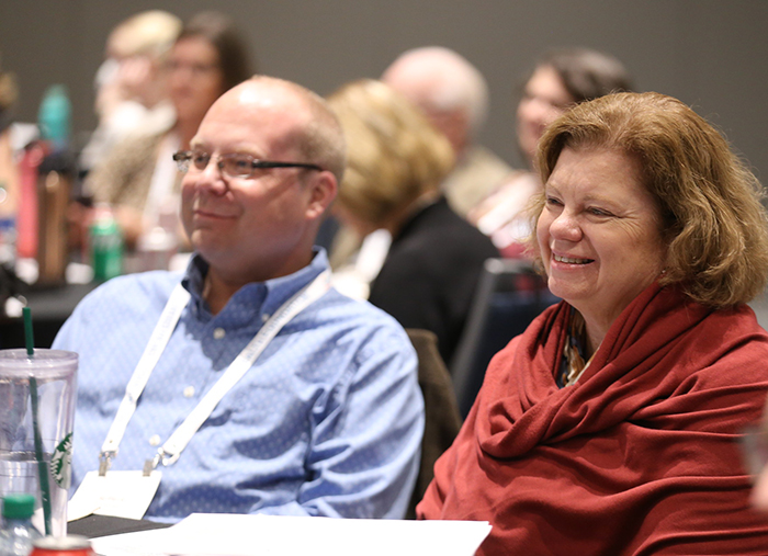 A white man and a white woman sit at a table and smile at the presenters during the Council of Affiliates meeting. 