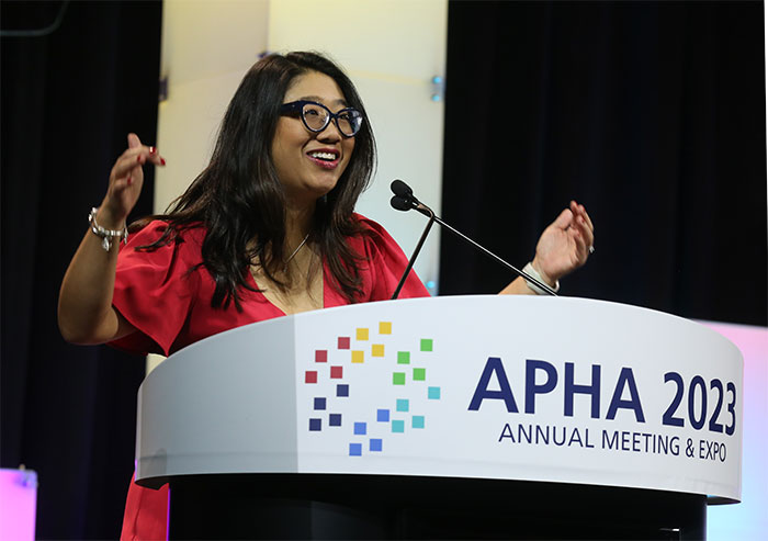 APHA President Chris Chanyasulkit smiles and gestures behind a podium at the APHA Opening Session. 