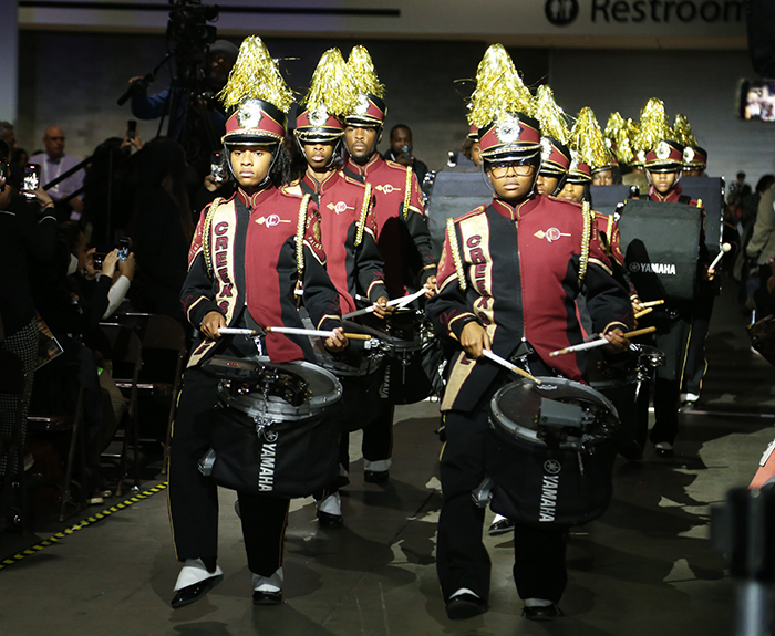 The front of the Creekside High School Band Drumline drums and marches down an aisle with APHA attendees standing and facing them on either side of the aisle. 