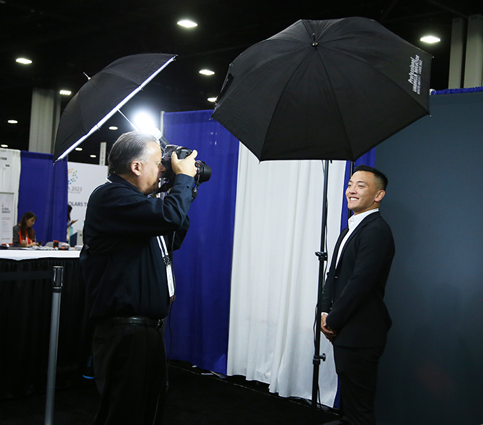 A young man stands proud and smiling against a black backdrop as a professional photographer takes his headshot at APHA Central in the exhibit hall. 