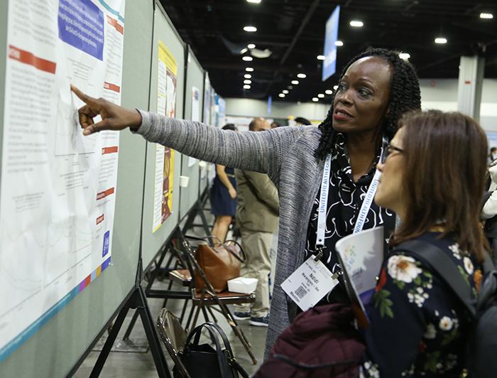 A Black woman with braids points to her poster in the APHA poster session as she explains her findings to another woman. 