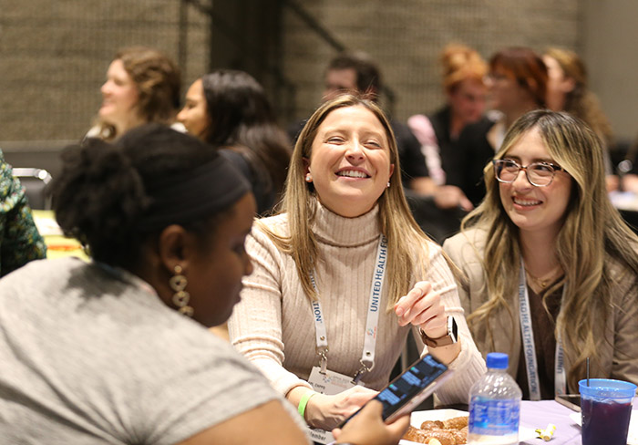 A young blonde woman laughs while closing her eyes and tilting her head back while another blonde woman sits next to her smiling; a Black woman sits sideways next to them. 