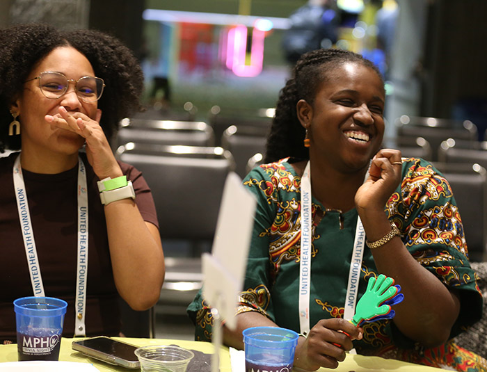 Two young Black women sit next to each other and laugh at something. One woman has her hand to her mouth while chuckling. 