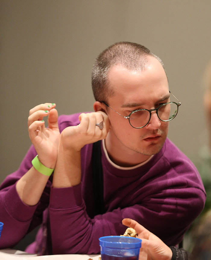 A person with a buzz cut and round glasses rests their elbows on the table while leaning over to look at something a person next to them is showing off camera. 