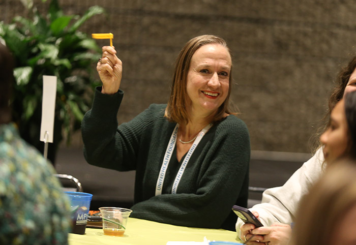 A smiling white woman with straight brown hair just below her shoulders raises a small plastic yellow flag during trivia night. 
