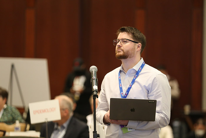 A person with a reddish-brown beard and brown hair holds an open laptop while standing in front of a microphone preparing to speak. 