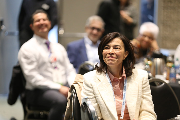 A middle-aged woman sits sideways in a chair smiling toward the front of the room at a session. 