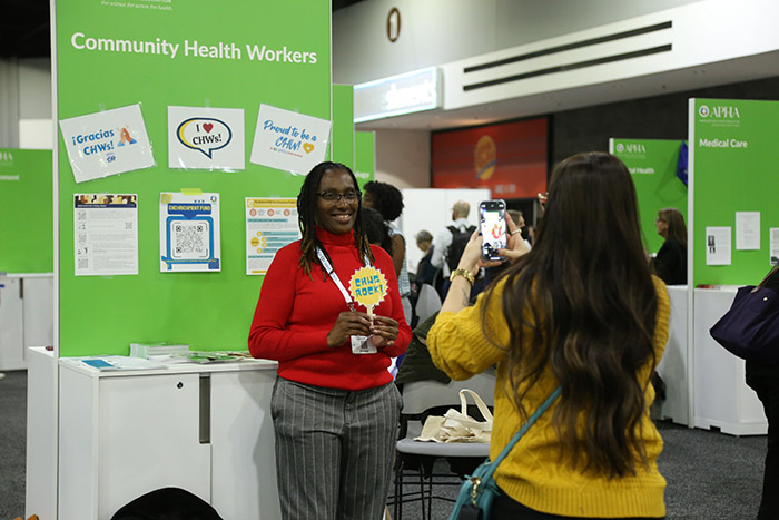 A Black woman in red sweater smiles and stands in front of the Community Health Workers booth while another woman takes a photo of her with a smartphone.