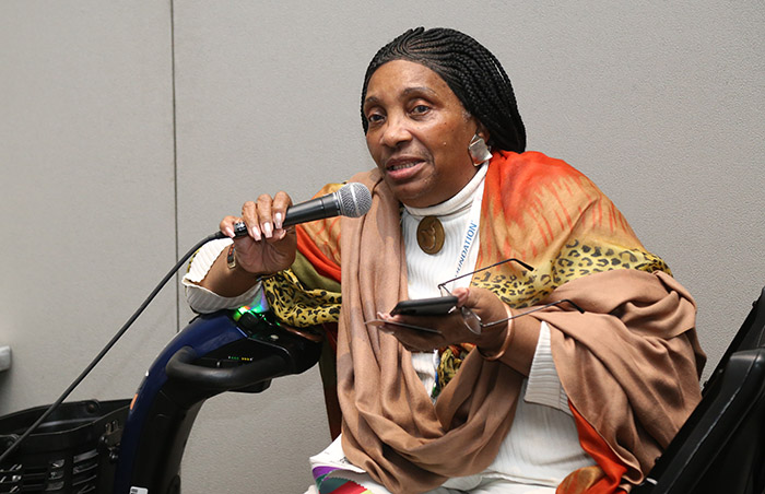 An older black woman wearing a multicolored scarf and sitting on a scooter holds a smartphone and eyeglasses in her left hand a microphone in her right hand while speaking. 