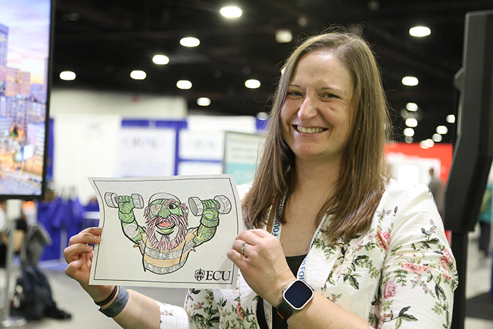A white woman grins at the camera while holding up a piece of paper with a picture on it that she colored. 