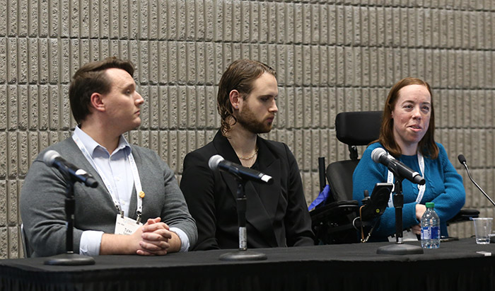 A woman in a blue shirt sits in a wheelchair while speaking in a session; next to her are two men who are fellow panelists. 