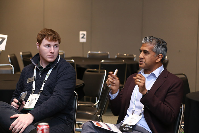 A man in a maroon blazer and blue shirt speaks while sitting in a chair during a session discussion; a younger man sitting next to him  watches and listens. 