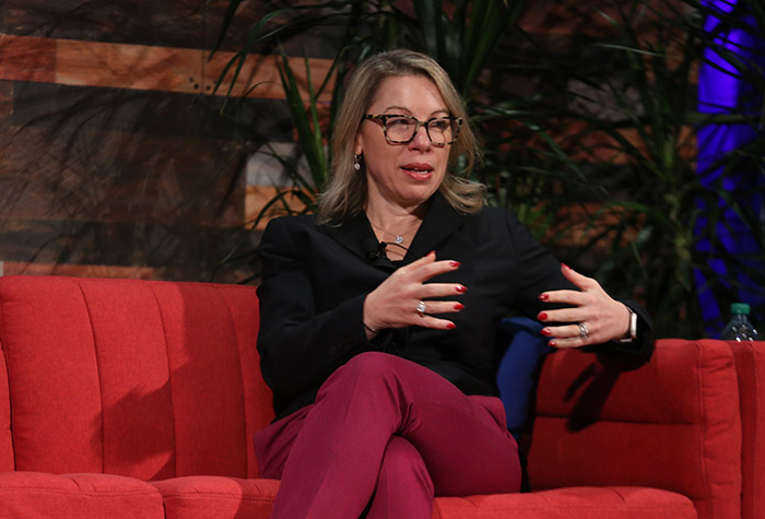 A blonde white woman wearing tortoise-shell eyeglasses, a black blouse and dark red pants sits with her legs crossed on a red couch on stage while gesturing with her hands while speaking. 