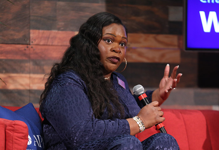 A Black woman with long black hair and wearing a purple outfit and gold hoop earrings holds a microphone and speaks on stage while sitting on a red couch. 